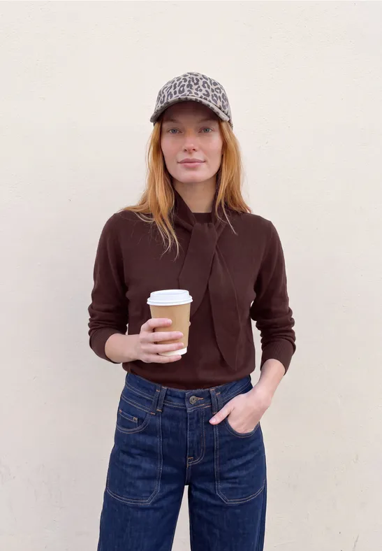 Eine junge Frau mit Cap und Kaffeebecher steht vor einer hellen Wand.