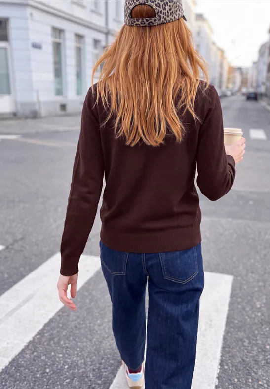 Eine Frau in Jeans, braunem Pullover und Leoparden-Cap steht auf einem Zebrastreifen und hält einen Kaffee zum Mitnehmen.