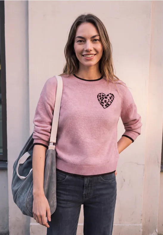 Eine lächelnde Frau in rosa Sweatshirt mit Herzprint trägt eine Tasche und lehnt an einer Hauswand.