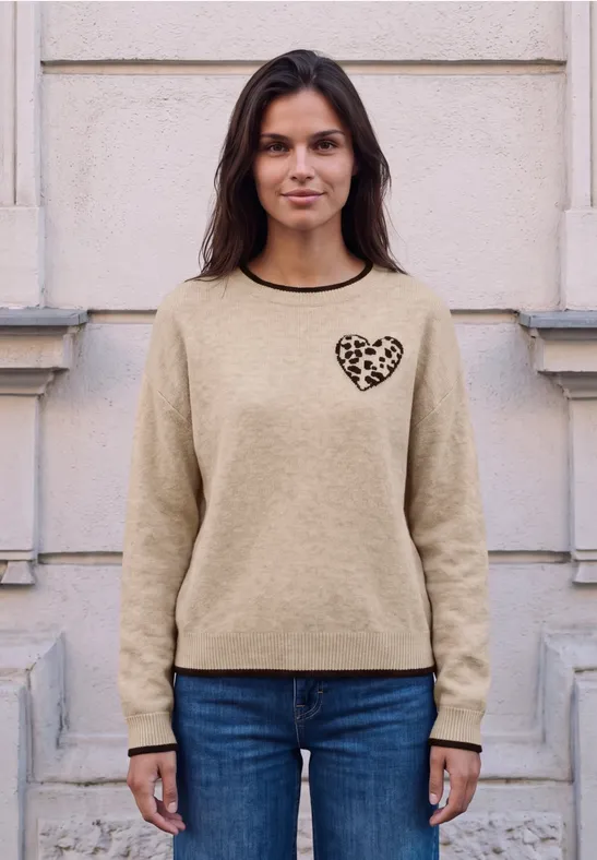 Frau steht vor heller Mauer, trägt beigen Pullover mit Leopardherz auf der Brust und blaue Jeans