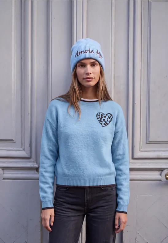 Eine Frau trägt eine hellblaue Mütze mit Schriftzug, einen blauen Pullover mit Herz und schwarze Jeans.