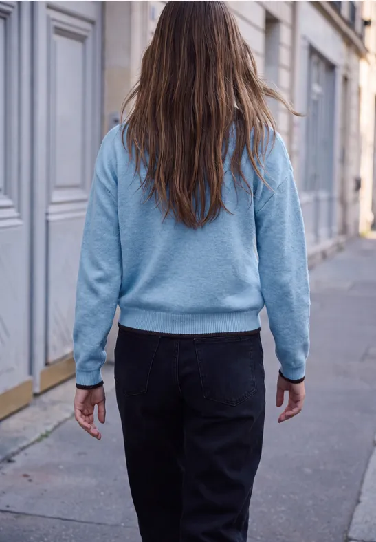 Rückenansicht einer Person mit langem Haar, hellblauem Pullover und dunkler Jeans auf einem Gehweg vor Häusern.