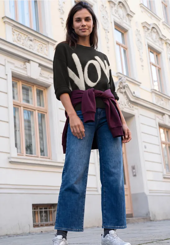 Eine junge Frau in Jeans und Pullover mit Aufdruck 'NOW' steht lässig vor einem Gebäude.
