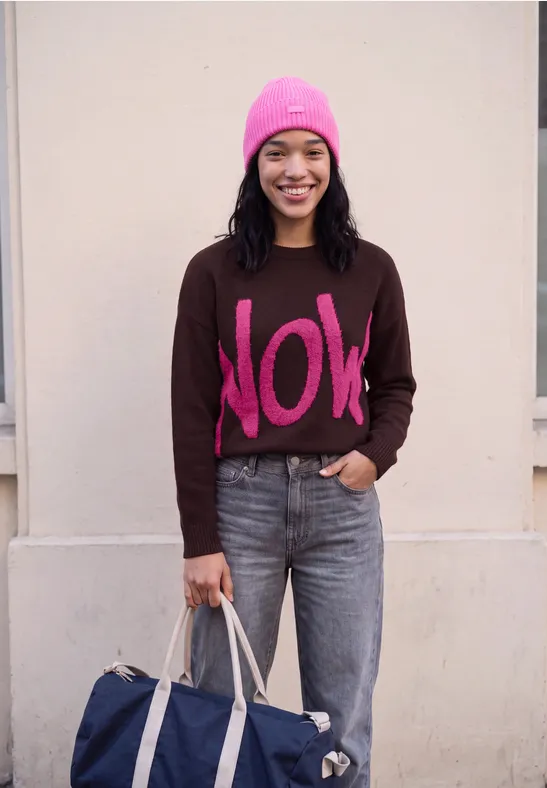 Lächelnde Frau mit pinker Mütze, braunem "NOW"-Pullover, Jeans und blauer Tasche vor neutraler Wand.