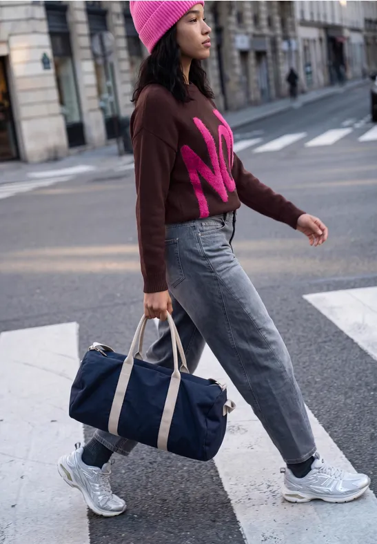 Frau mit rosa Mütze und Pullover überquert Straße, trägt blaue Reisetasche und Sneakers.