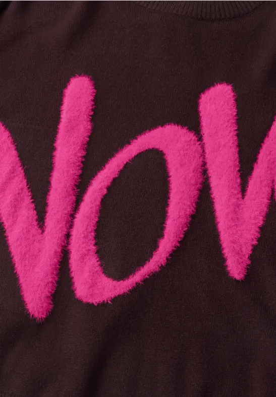 Brauner Pullover mit flauschigen, pinken Buchstaben "VOV" auf Brusthöhe.