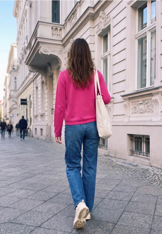 Eine Frau in pinkem Pullover, Jeans und weißen Sneakern geht die Straße entlang, trägt eine weiße Tasche.