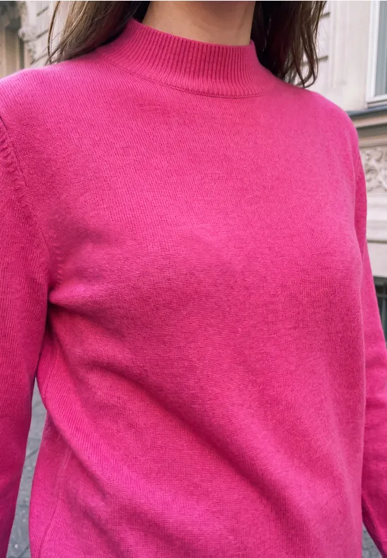 Eine Frau trägt einen pinkfarbenen Pullover vor einer Hauswand.