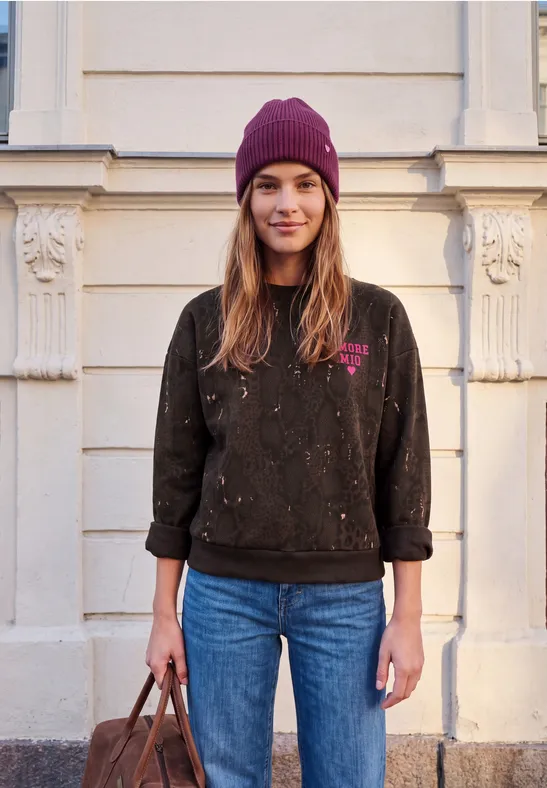 Junge Frau mit lila Mütze, schwarzem Pullover und Jeans steht vor hellem Gebäude und hält eine Tasche.