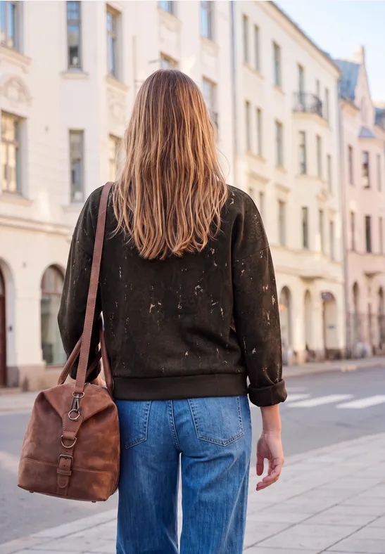 Frau mit brauner Ledertasche, schwarzem Pullover und Jeans geht eine Stadtstraße entlang, gesehen von hinten.