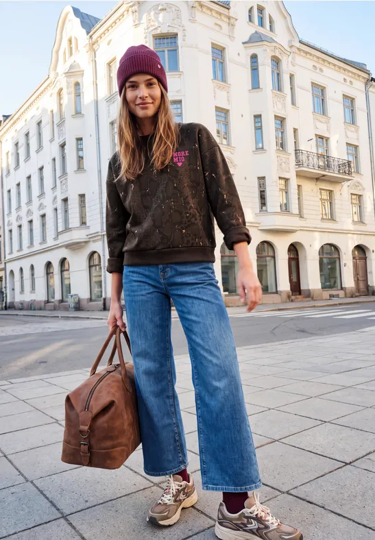 Junge Frau mit lila Mütze, weiter Jeans und Metallic-Sneakern posiert vor weißem Altbau, hält Reisetasche.