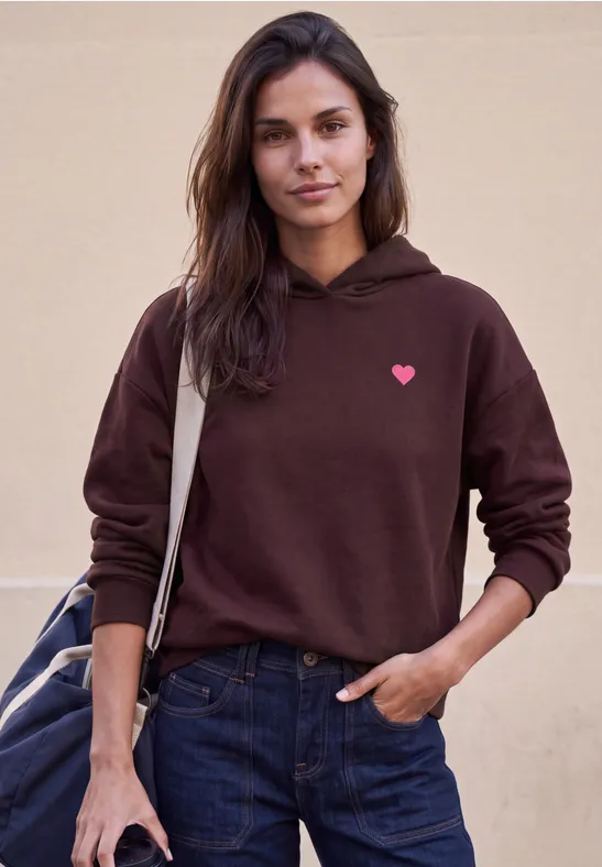 Eine Frau in braunem Hoodie mit kleinem rosa Herz, Jeans und Tasche.