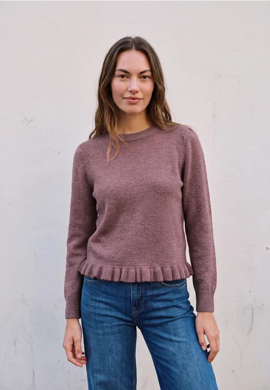 Eine junge Frau trägt einen mauvefarbenen Pullover und Jeans vor hellgrauer Wand.