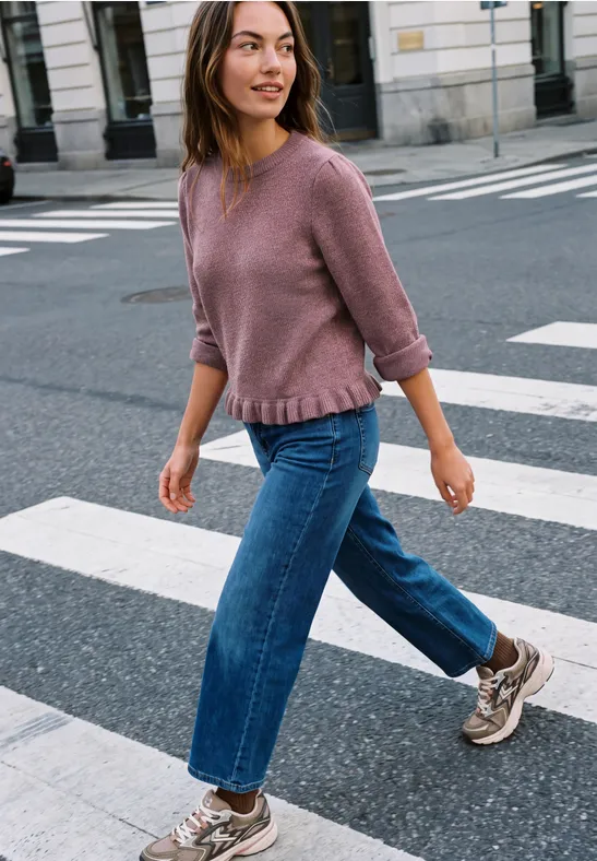 Eine Frau in rosa Pullover, blauen Jeans und Sneakern überquert lächelnd einen Zebrastreifen.
