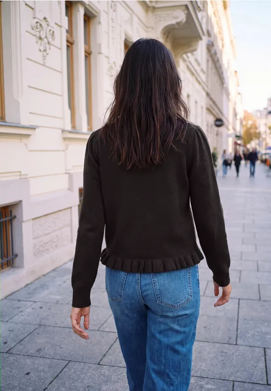Person mit langen, dunklen Haaren von hinten, in schwarzer Jacke und blauen Jeans auf einer städtischen Straße.