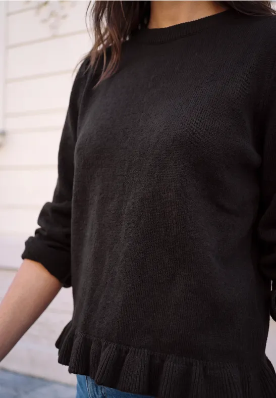 Schwarzer Strickpullover mit Rüschen am Saum, getragen von Frau in heller Umgebung.