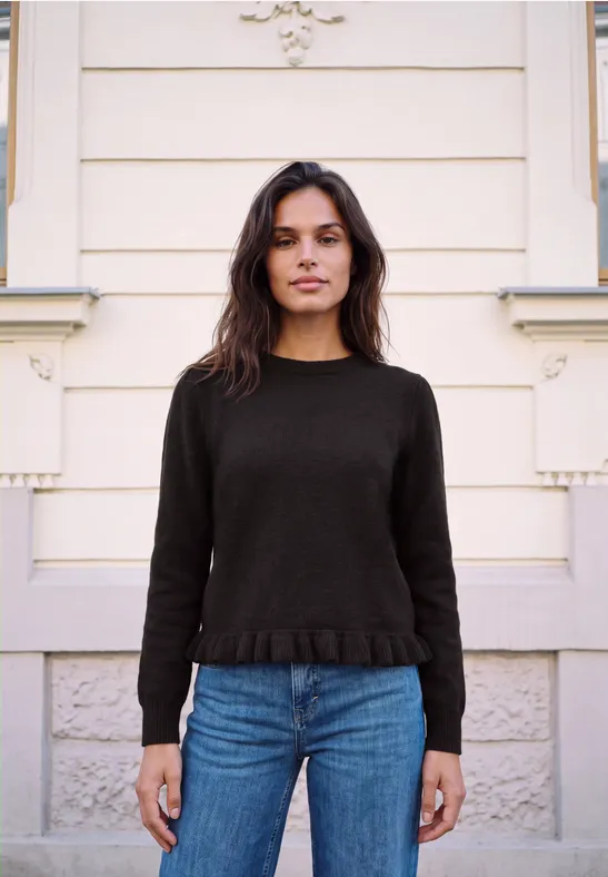Junge Frau vor heller Hausfassade, trägt schwarzen Pullover und blaue Jeans, schaut in die Kamera.