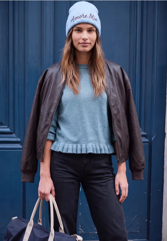 Junge Frau mit grauer Mütze, brauner Lederjacke, grauem Pullover, schwarzen Jeans und Tasche vor blauem Tor.