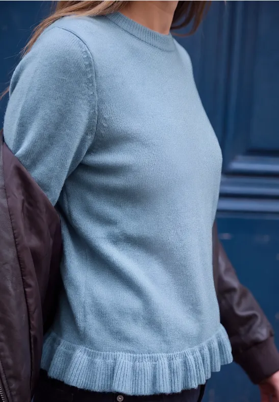 Trägt hellblauen Pullover mit Rüschenbund und Lederjacken-Ärmeln vor dunkelblauem Hintergrund.