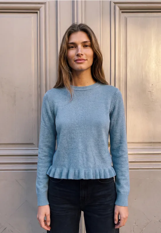 Eine Frau im blauen Pullover steht vor einer hellen Wand und blickt in die Kamera.