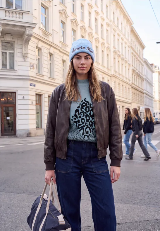 Junge Frau mit Jeans, Lederjacke und Leo-Pullover steht in Straßenszenerie.