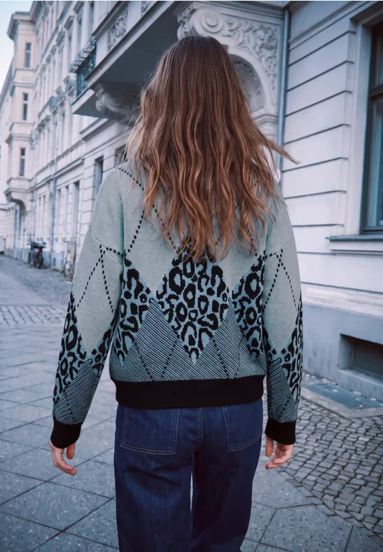 Eine Person in grauem, gemustertem Pullover und Jeans geht eine Straße entlang, von hinten aufgenommen.
