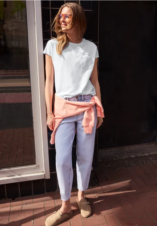 Person steht vor Gebäude, trägt weißes T-Shirt, hellblaue Jeans, braune Schuhe und rosa Pullover um die Taille.