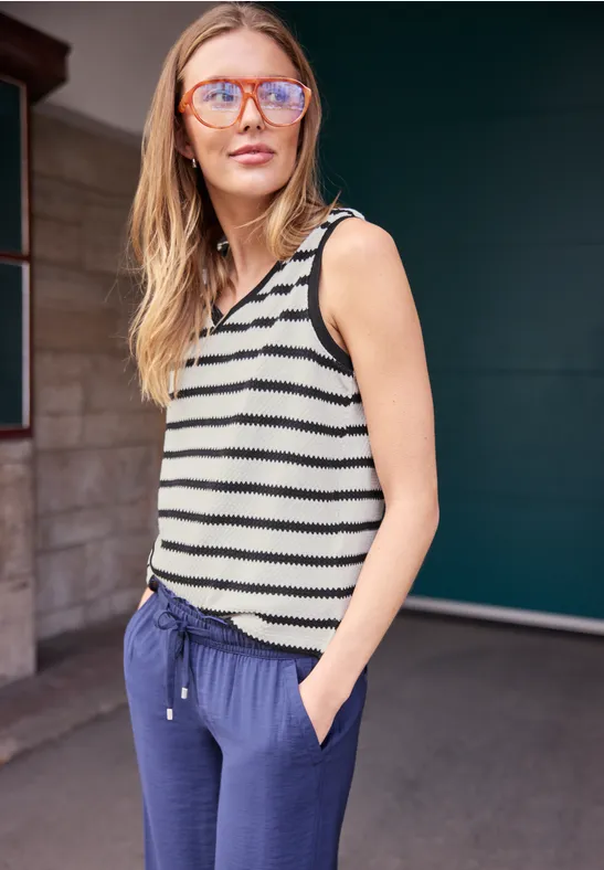 Eine Frau im gestreiften Top und blauer Hose steht lässig mit Händen in den Taschen.