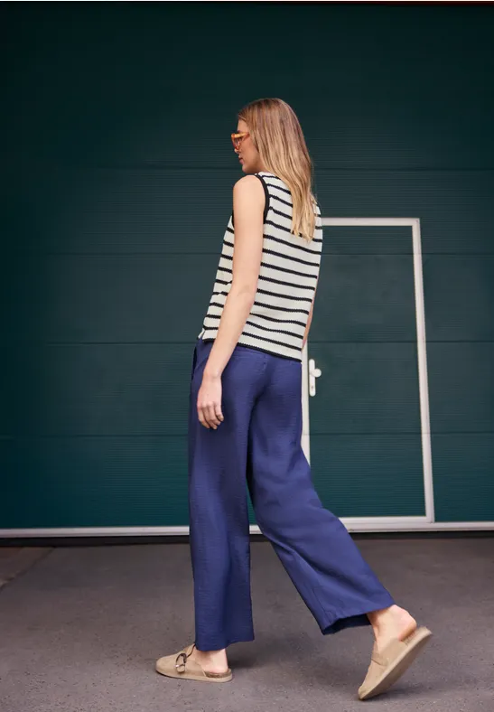 Eine Frau in gestreiftem Top und weiten blauen Hosen läuft seitlich vor einer dunklen Wand mit Tür.