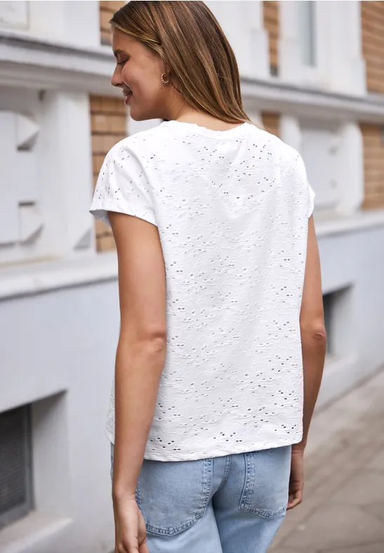 Een vrouw van achteren in een wit ajourshirt en blauwe spijkerbroek op een stoep.