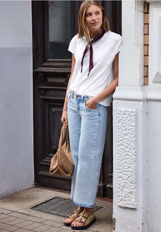 Persoon staat bij een gebouwingang, draagt een witte blouse, lichtblauwe spijkerbroek, sjaal, sandalen en bruine tas.