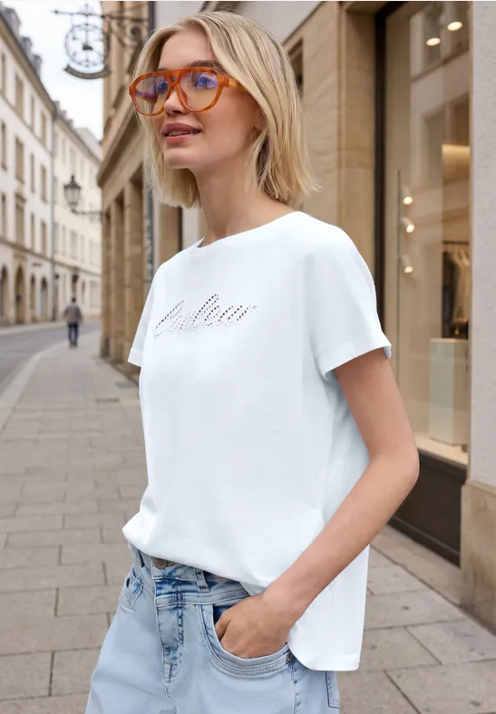 Eine Frau in weißem T-Shirt und Jeans steht mit der Hand in der Hosentasche auf einer Stadtstraße.