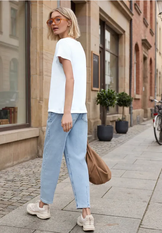 Frau in weißem T-Shirt, hellen Jeans und Sneakers, trägt Tasche, spaziert an einer Hausfassade entlang.