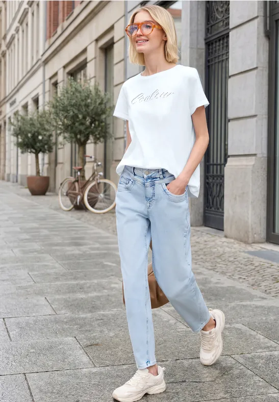 Junge Frau in Jeans, weißem T-Shirt und Sneakers geht entspannt über eine Stadtstraße.