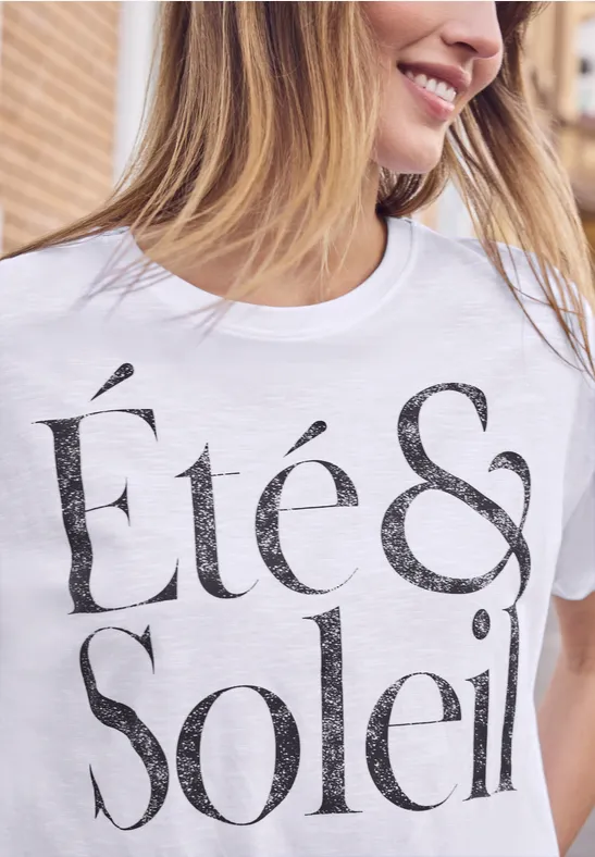 Nahaufnahme einer lächelnden Frau im weißen T-Shirt mit dem Schriftzug „Été & Soleil“.
