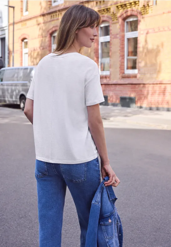 Eine Frau steht in Jeans und weißem T-Shirt auf der Straße und hält eine Jeansjacke in der Hand.