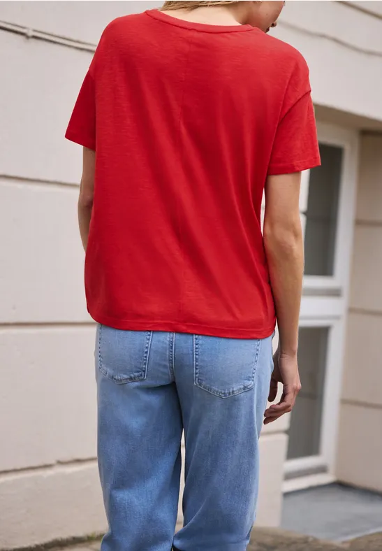 Achteraanzicht van een persoon in een rood T-shirt en blauwe jeans voor een witte gevel met deur.