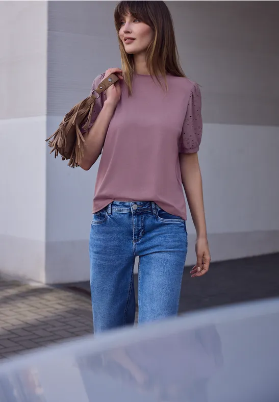 Frau mit rosa Oberteil, blauer Jeans und brauner Fransen-Tasche läuft draußen.
