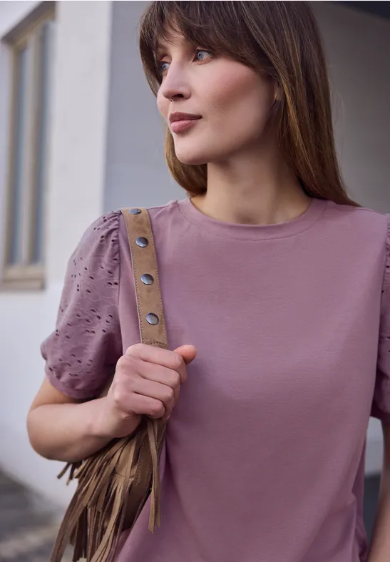 Junge Frau steht vor Gebäude, trägt hellviolettes T-Shirt mit Nietenbesatz und Fransen-Umhängetasche.