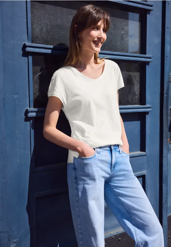 Jonge vrouw in wit T-shirt en blauwe jeans, leunend tegen een blauwe deur, handen in de zakken.