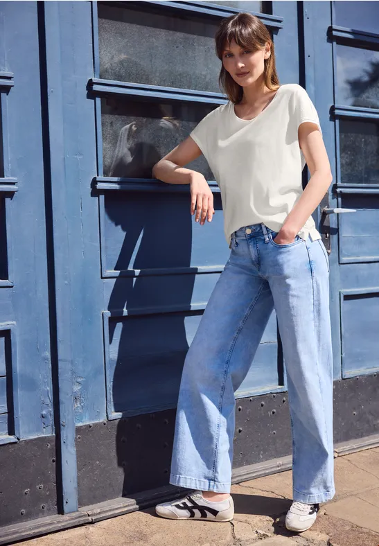 Een vrouw in wit T-shirt en losse jeans leunt nonchalant tegen een blauwe metalen deur buiten.