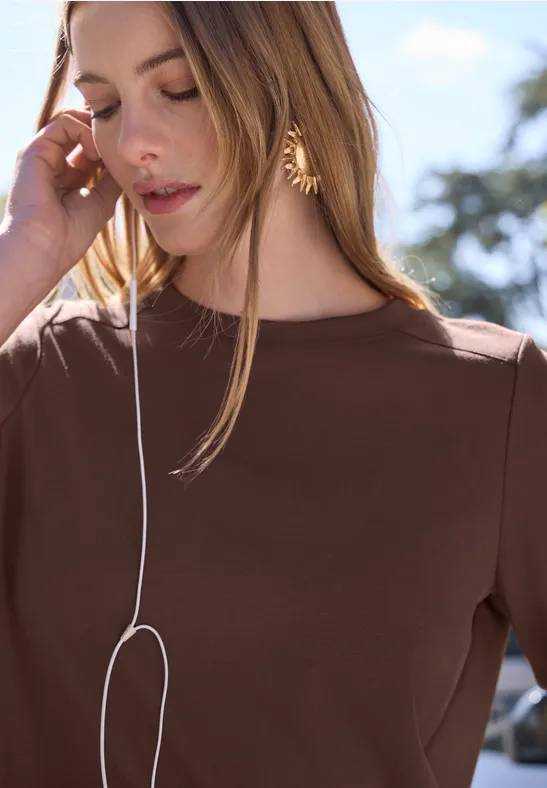 Femme en haut marron et cheveux longs, en extérieur, parlant au smartphone avec des écouteurs blancs.