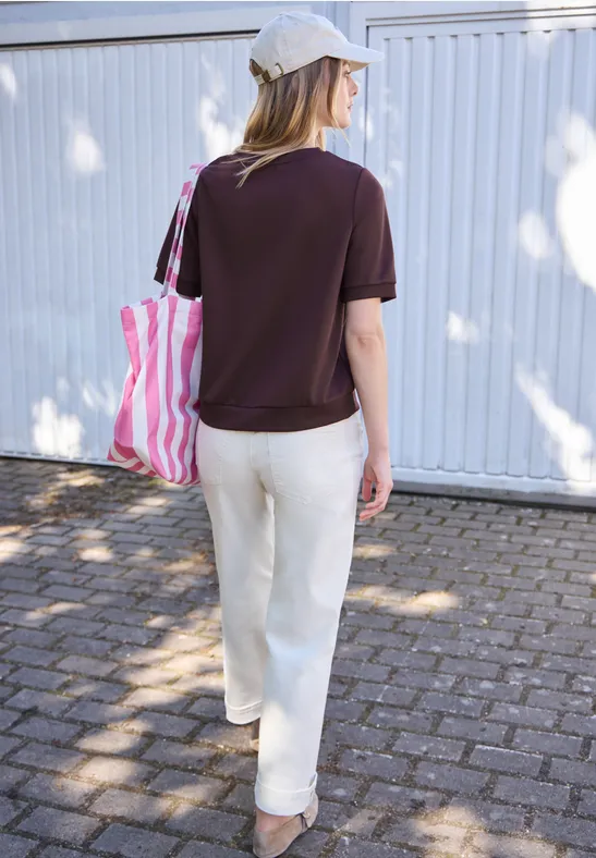 Frau von hinten mit weißer Kappe, dunkelrotem Shirt, weißer Hose und rosa-weiß gestreifter Tasche vor weißer Wand.