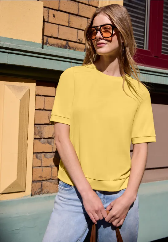 Femme en chemise jaune et jean, appuyée de manière décontractée contre un mur en brique.