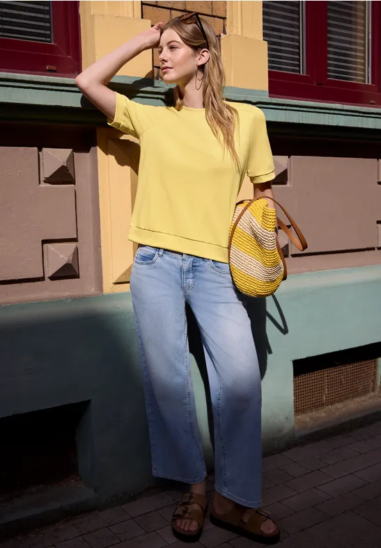 Femme en t-shirt jaune, jean large bleu clair et sac en paille, se protège du soleil d’une main en ville.