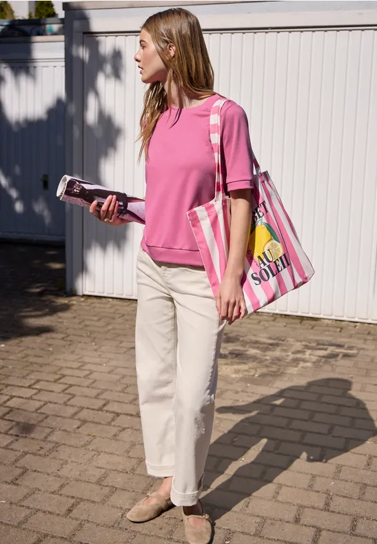 Vrouw in roze shirt en witte broek voor een wit hek, met telefoon in de hand en kleurrijke tas.