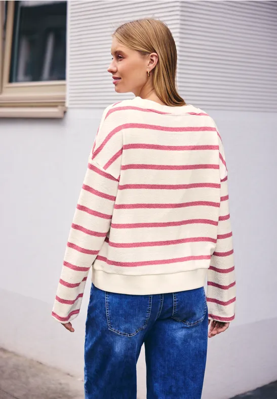Eine Person steht vor einer Wand, trägt einen gestreiften Pullover und blaue Jeans.