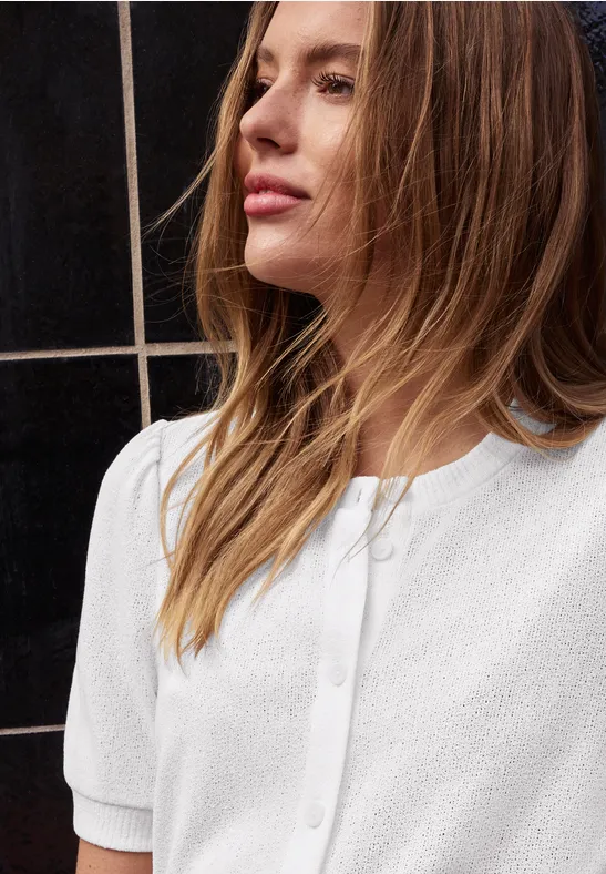 Personne aux cheveux blond-castanés longs porte un chemisier blanc à manches courtes devant un carrelage noir.