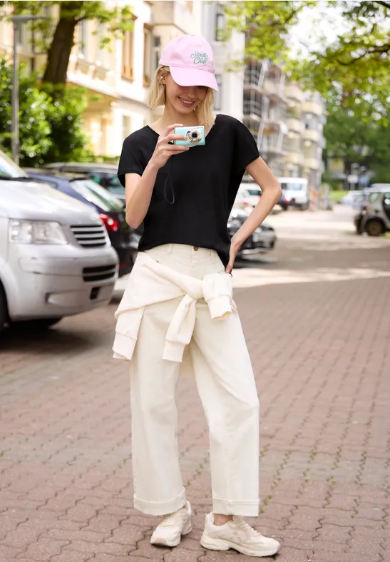 Junge Frau mit rosa Cap, schwarzem Shirt und weitem weißen Hose trinkt Kaffee auf Gehweg vor parkenden Autos.