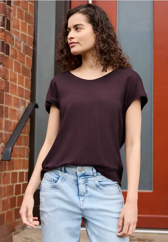 Junge Frau mit dunklem T-Shirt und heller Jeans steht vor einer Tür und orangener Backsteinwand.